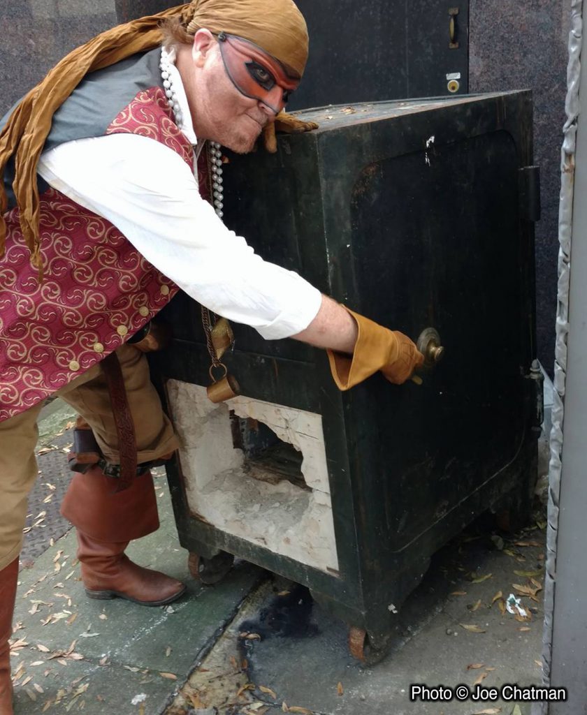 Captain Drew wrestles with a giant old-fashioned safe on the sidewalk. The side of the safe appears to have been blown open.